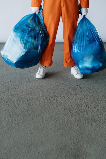 A person standing on a grey concrete floor is partially visible from the waist down, wearing orange coveralls and white sneakers, holding two large blue plastic bags filled with waste, with the bags slightly transparent revealing various discarded items inside. The background is a plain light-colored wall, and the scene suggests an activity related to waste collection or rubbish disposal, consistent with professional waste management services such as those offered by Waste Clearance Bayswater for private rubbish removal or on-site clearance solutions.