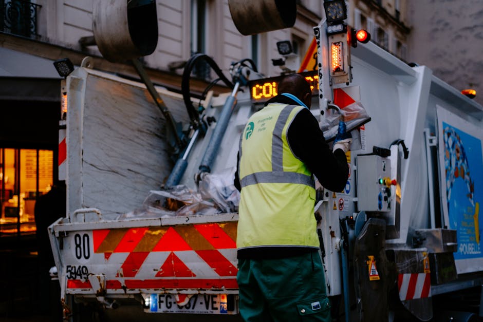 A worker wearing a high-visibility yellow vest with reflective stripes and dark clothing operates a waste collection vehicle on a city street, with buildings and illuminated windows in the background. The vehicle is equipped with a mechanical arm and hydraulic system for loading rubbish, and the rear of the truck is partially open, revealing a load of mixed waste materials including paper and plastic debris. The scene is set during dusk or early evening, with ambient lighting casting a soft glow. The waste management process involves the collection and unloading of waste, typical of independent or private rubbish removal services like those offered by Waste Clearance Bayswater. The environment is urban, with a focus on efficient rubbish collection for disposal or transfer to waste facilities.