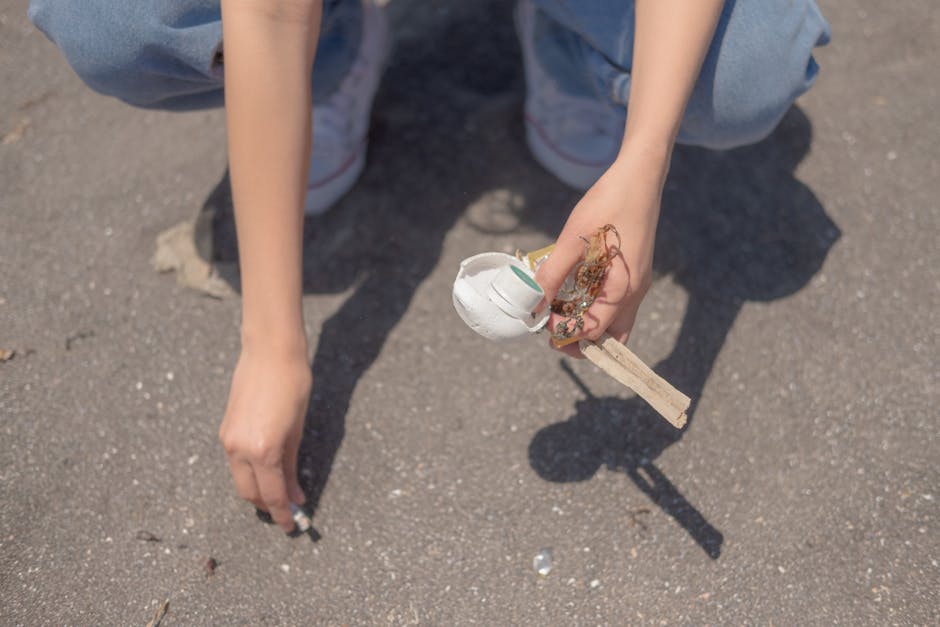 A person is crouching on a paved outdoor surface, holding a small wooden stick with a broken, partially painted white egg shell attached to it. The egg shell has some visible dirt and remnants of eggshell texture. The individual is wearing light blue jeans and white sneakers with red and blue accents. Their arm and hand are visible but their face and upper body are not shown. The background consists of a flat, gray, textured pavement with small scattered debris. The scene highlights a moment of casual littering or collection, which relates to disposal and waste management practices. Waste Clearance Bayswater’s rubbish removal services support proper disposal of discarded items like broken eggs or similar waste found in outdoor environments, emphasizing environmentally responsible clearance methods.