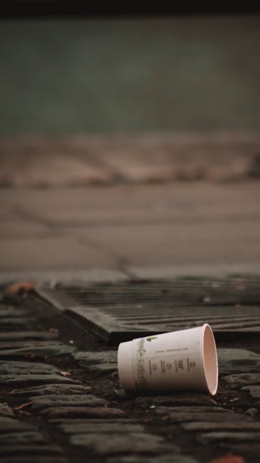 A white paper cup with green printed text lies on a cobbled surface next to a metal grate, with a blurred background of paving slabs and a concrete curb. The cup appears discarded, and the surrounding environment suggests an outdoor urban setting, possibly a street or pavement area. The surface textures include the rough, uneven cobblestones and the metal grate's ridged pattern. The scene is lit with natural, diffuse lighting, creating subtle shadows. This image reflects the common issue of littering in public spaces and highlights the importance of proper rubbish disposal, which Waste Clearance Bayswater can assist with through alternative waste handling and private rubbish removal services in the local area.