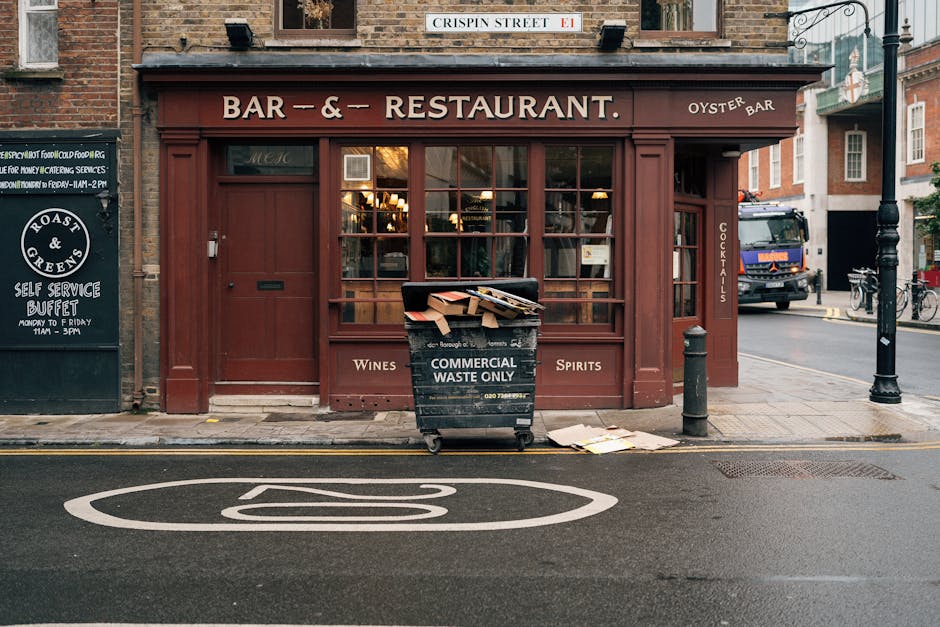 In front of a traditional brick-fronted bar and restaurant with large window panes showcasing warm interior lighting, a large black commercial waste bin marked 'Commercial Waste Only' stands on the pavement. The bin is overflowing with flattened cardboard boxes and paper waste, some of which are spilling onto the ground nearby. To the right of the bin, a black bollard and a metal street lamp post are positioned along a narrow city street with marked parking restrictions painted on the asphalt. The scene is set during daylight, with natural ambient lighting highlighting the textures of the red-brown bar facade, the corrugated cardboard, and the surrounding urban environment. This setup indicates an alternate approach to waste disposal and rubbish removal services for businesses, such as private collection by Waste Clearance Bayswater, ensuring the collection of commercial waste in a tidy manner outside the premises.