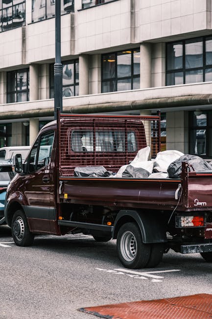 A small burgundy flatbed truck parked on a city street in front of a modern building with large glass windows and concrete facade. The truck's cargo bed is filled with various types of waste materials, including large black and white bags, some open and showing contents, as well as several loose objects. The truck features a metal mesh guard at the front of the cargo area to prevent items from falling out during transit. Its rear tailgate is down, and the vehicle is positioned close to the curb, suggesting it is engaged in rubbish removal or on-site clearance. The surroundings include other parked vehicles, a lamppost nearby, and a clearly marked parking bay, all within an urban environment that may involve private waste collection services such as those offered by Waste Clearance Bayswater for alternative disposal options outside local authority collections.